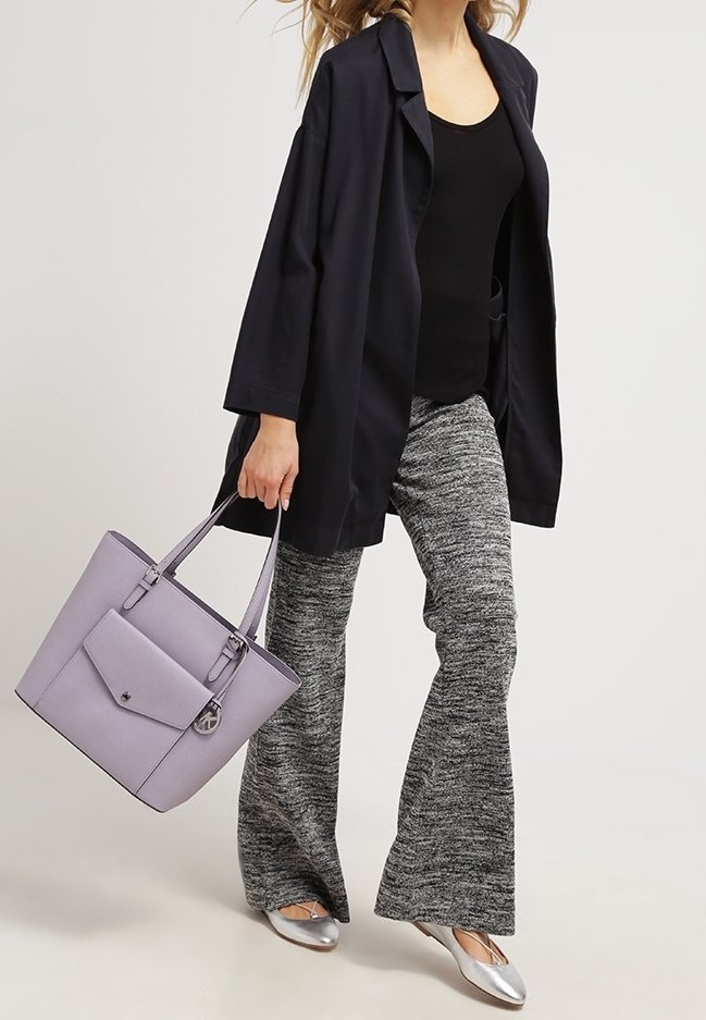 Personne portant un haut noir et une veste, un pantalon gris texturé, des ballerines argentées, portant un sac cabas violet clair avec une poche avant et un porte-clés.