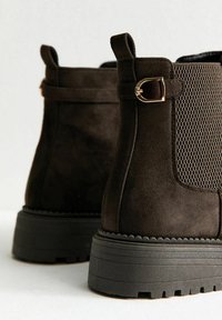 Een paar donkerbruine suède enkelschoenen met chunky zwarte zolen, elastische patroon zijpanelen en kleine gouden gespen op de achterbanden.