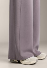 Široké, levandulové tepláky vyrobené z jemného materiálu s žebrovanými bočními panely, v kombinaci se světle zbarvenými teniskami s texturovanými prvky.