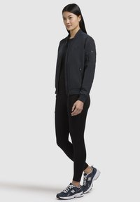 Giacca bomber blu navy con zip frontale, polsini a coste e tasca laterale. Abbinata a leggings neri e sneakers blu navy di New Balance.