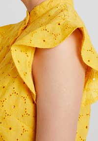 Yellow floral-embroidered blouse featuring a high collar and ruffled sleeves. The fabric has a textured, lightweight appearance.