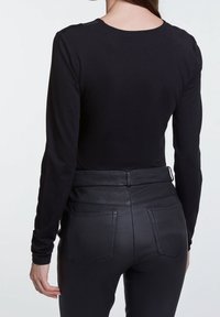 Vrouw in een aansluitend zwart shirt met lange mouwen en een hoge taille van zwarte leren broeken, gezien van achteren tegen een effen achtergrond.