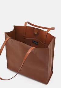 Sac fourre-tout en cuir marron avec deux poignées supérieures, structure ouverte et fermeture à pression. Présente une texture lisse et une petite poche intérieure.