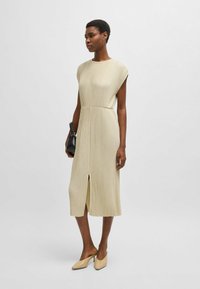 Beige, knee-length dress with vertical pleats, cap sleeves, and a front slit. Paired with nude mules and a small black handbag.