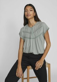 Jasnozielona bluzka z okrągłym dekoltem, krótkimi rękawami i dekoracyjnym przeszyciem. Luźny krój z marszczonym wykończeniem w talii.