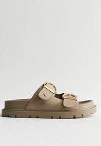 Brun slide sandal gjord av slätt material, med två gyllene spännen, en tjock sula med ränder och en elegant, minimalistisk design.