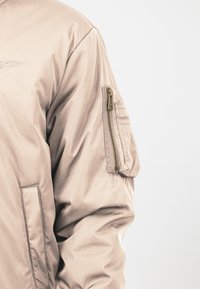 Beżowa kurtka bomberka z zamkniętą kieszenią na rękawie, gładką teksturą i lekko wyściełanym fasonem z subtelnymi detalami w szwach.