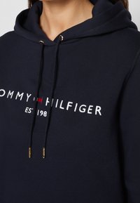 Felpa blu navy con coulisse e punte dorate, caratterizzata da ricamo con la scritta "Tommy Hilfiger EST 1985" e logo rosso, bianco e blu.