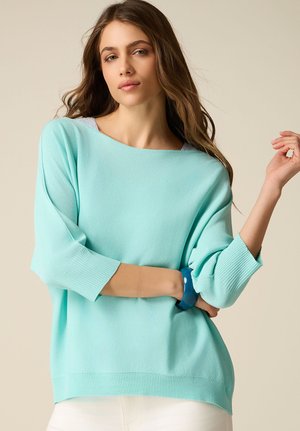 Jonge vrouw die een losse muntgroene trui en witte broek draagt, poseert met één arm gekruist en een blauw armbandje om haar pols.