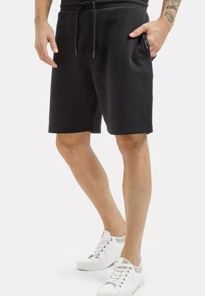 Osoba ubrana w czarne spodenki ze sznurkiem, ręce blisko kieszeni, widoczne tatuaże na nogach oraz białe sneakersy sznurowane.