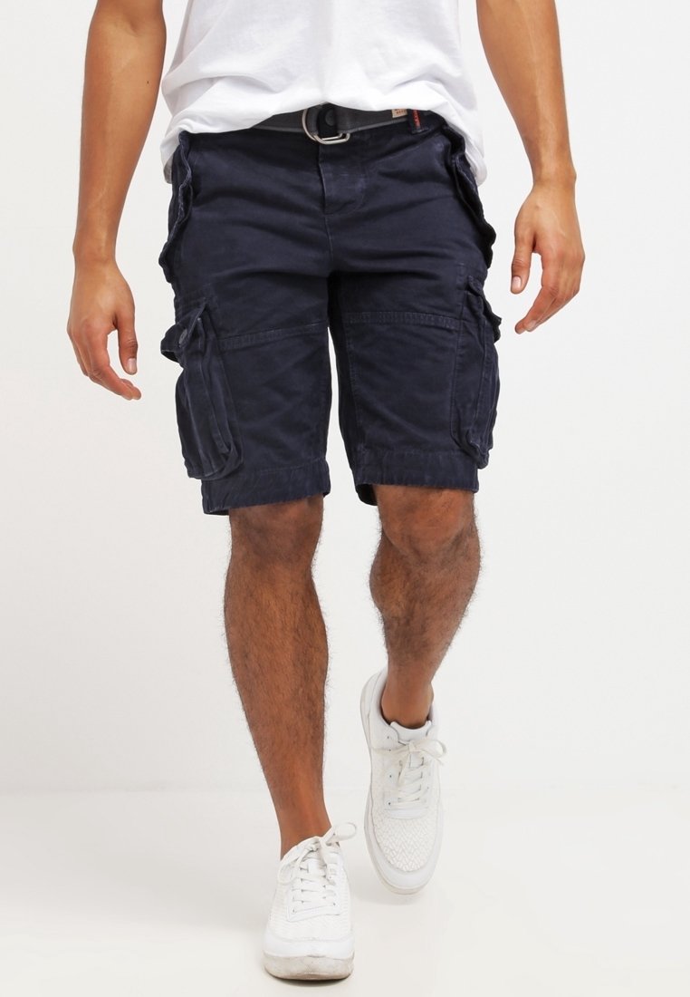 Homme portant un short cargo bleu foncé, des baskets blanches et un t-shirt blanc marchant vers l'avant sur un fond blanc uni.