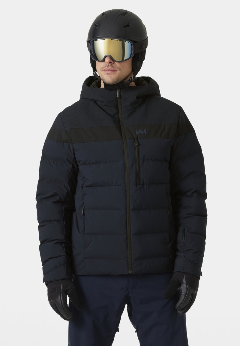 Marineblaues Steppjacke mit einem schwarzen oberen Panel, Reißverschluss vorne und elastischen Bündchen. Trägt einen schwarzen Skihelm und verspiegelt Sonnenbrille. Schwarze Handschuhe.