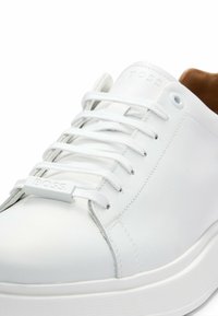 Basket en cuir blanc avec lacets blancs, détail embossé "BOSS" sur l'étiquette des lacets et doublure intérieure marron visible au talon.
