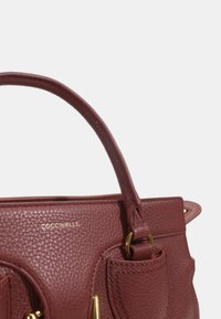 Borsa a tracolla in pelle bordeaux con texture, manico superiore, dettagli dorati, tasca frontale e logo impresso. Forma compatta con design strutturato.