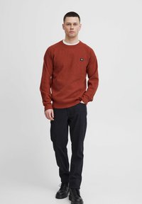 Maglione a maglia color ruggine con scollo rotondo e un piccolo etichetta del marchio. Abbinato a pantaloni scuri e stivali neri con lacci.