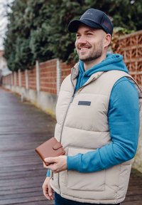 Beige puffer vest over a teal hoodie, holding a brown leather wallet. The setting features wooden planks and greenery in the background.