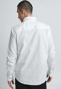 Chemise blanche à manches longues avec une texture douce et des poignets à boutons. Elle présente un col évasé, un ourlet arrondi et une couture de carreau au dos.
