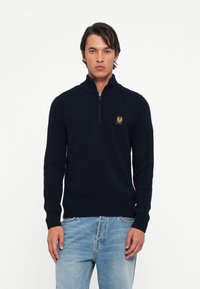 Pulover tricotat cu fermoar de culoare bleumarin, cu guler înalt, manșete și tiv cu nervuri, având un patch cu logo pe piept și dungi verticale texturate pe mâneci.