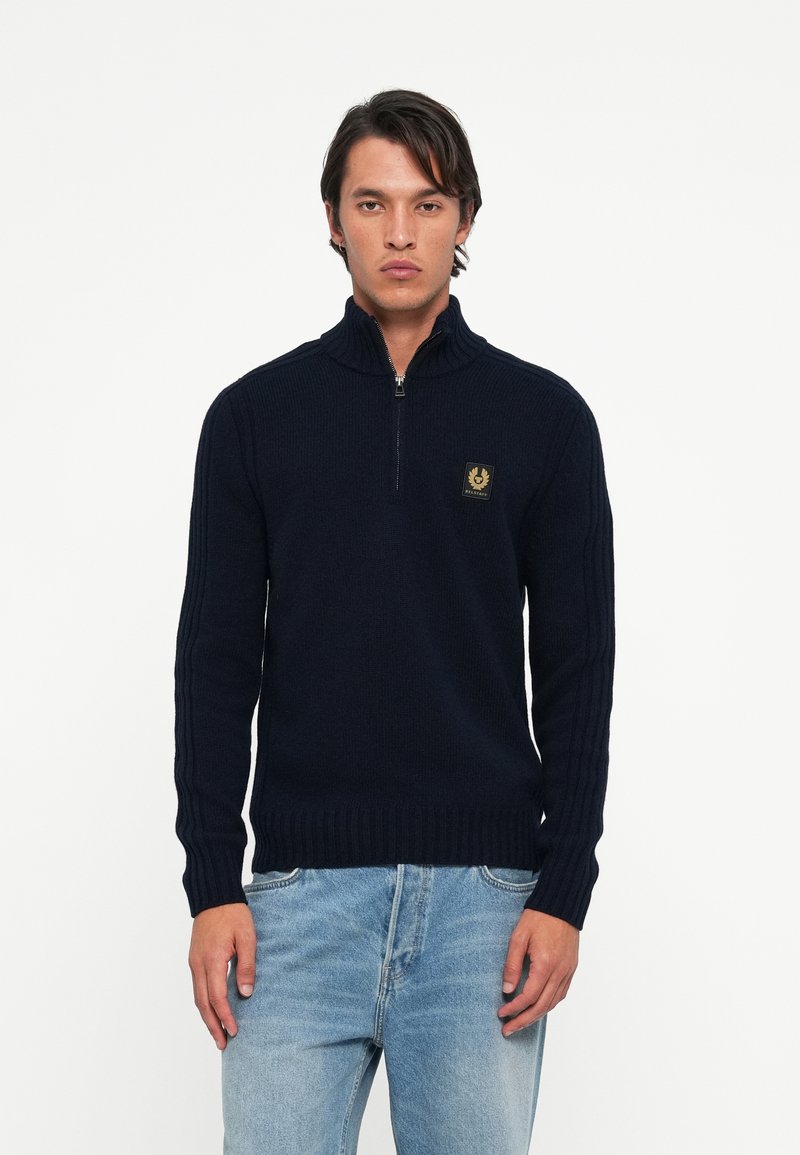 Pulover tricotat cu fermoar de culoare bleumarin, cu guler înalt, manșete și tiv cu nervuri, având un patch cu logo pe piept și dungi verticale texturate pe mâneci.
