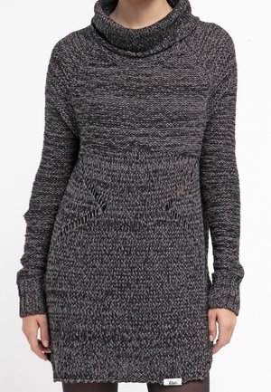 Kobieta ma na sobie długą, ciemnoszarą, grubą dzianinową sukienkę z golfem i detalami w postaci kieszeni z przodu.