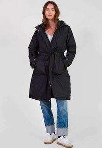 Zwart gewatteerde lange jas met een trekkoord in de taille, capuchon en drukknoopsluiting. Gecombineerd met lichtblauwe spijkerbroek en witte sneakers.