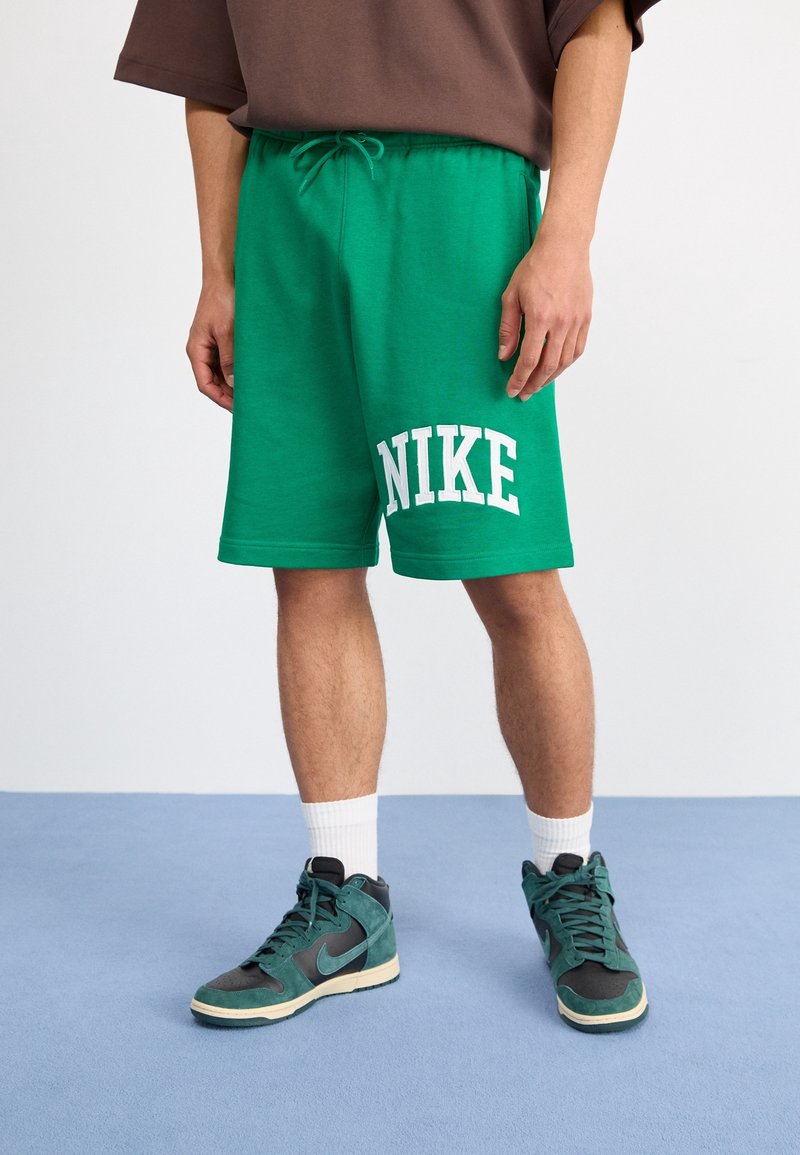 Green sweat shorts with white "NIKE" logo, elastic waistband, and drawstring. Worn with dark green high-top sneakers and white socks.