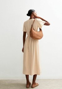 Beige midi-jurk met korte mouwen, vastgemaakt met een strik in de taille. Het model draagt een bruine leren hobo tas. Zanderige vloer en een lichte achtergrond.