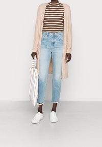 Beige lång kofta över en brun och vit randig skjorta, ljusblå högmidjade jeans och vita sneakers; håller en randig tote-väska.