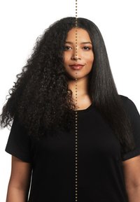 De helft van het haar van het model is volumineus en krullend, terwijl de andere helft steil en glad is. Ze draagt een zwarte shirt en poseert tegen een witte achtergrond.