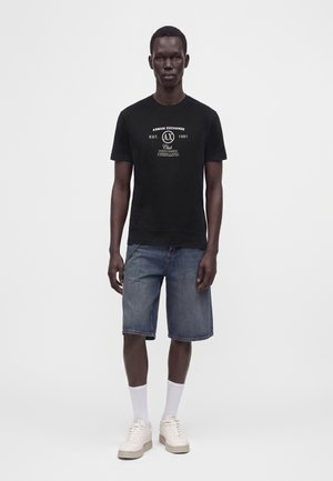 Homme portant un t-shirt noir Armani Exchange, un short en jean bleu, des chaussettes blanches et des baskets beige, debout devant un fond uni.