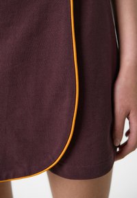 Burgundi színű ruha textúrált anyaggal és kontrasztos narancssárga paszpóllal az alján, sima felülettel és letisztult vonalakkal.