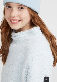 Pull en polaire bleu clair, doux et pelucheux, avec un col montant. Présente un logo en patch foncé sur la manche. Le modèle porte un bonnet en tricot assorti bleu clair.