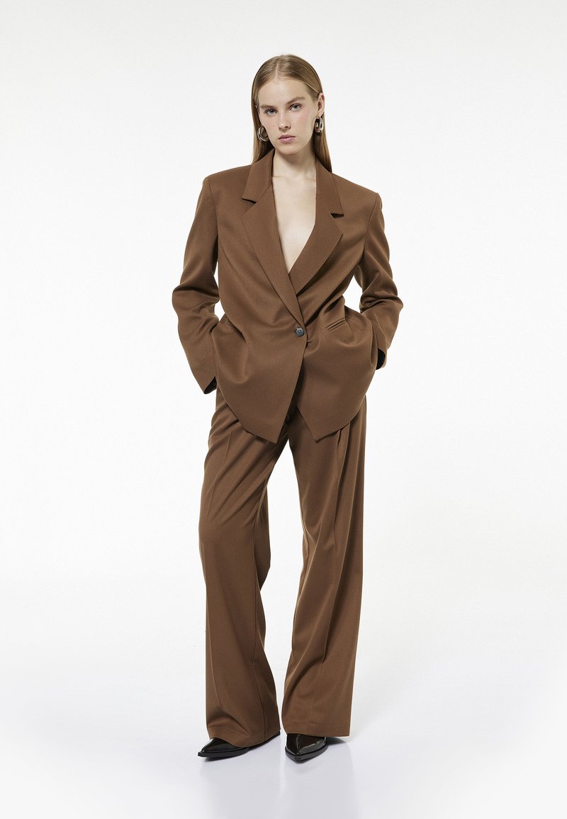 Brauner maßgeschneiderter Blazer mit Reverskragen und einer einzigen Knopfleiste, kombiniert mit locker sitzenden braunen Hosen. Schwarze spitze Pumps runden den Look ab.