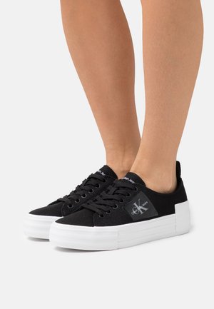 Svarta dunsneakers med vita platåformade sulor. Har rundad tå, snörning och logotypetikett på sidan. Texturerad yta.