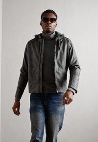 Graue Lederjacke mit Kapuze, Frontreißverschluss und gerippten Akzenten, über einem dunklen Rollkragenpullover getragen und kombiniert mit blauen Jeans.
