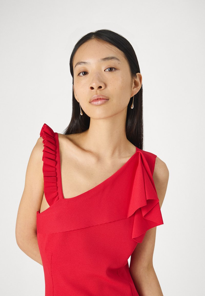 Czerwona asymetryczna sukienka z falbaniastymi akcentami na lewym ramieniu, wykonana z gładkiego materiału. Modelka nosi proste kolczyki wiszące dla kontrastu.