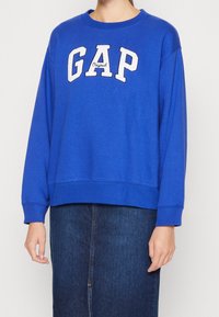 Sudadera azul con el texto "GAP" en blanco y una textura suave. Manga larga y corte relajado, combinada con una falda de mezclilla oscura.
