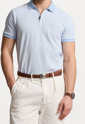 Jasnoniebieska koszulka polo w paski z zamkiem przy kołnierzu, zestawiona z beżowymi spodniami z plisami i brązowym skórzanym paskiem. Zegarek na lewej ręce.