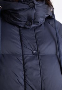 Marineblaue Steppjacke mit hohem Kragen, ausgestattet mit Knopfverschlüssen und Kordelzügen. Der Stoff ist glatt mit einem gesteppten Muster.