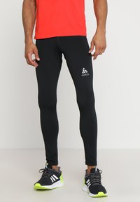Schwarze Sportleggins mit einer engen Passform, versehen mit einem weißen Logo am linken Bein und Reißverschlussakzenten am Knöchel, kombiniert mit einem roten Oberteil und schwarzen Schuhen.