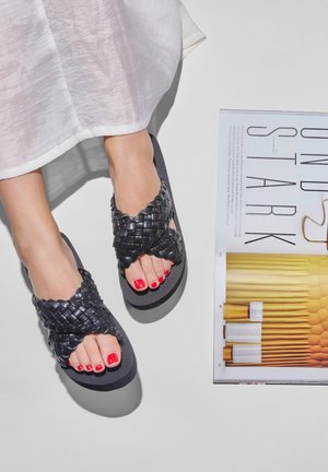 Black woven sandals with a textured strap design. The foot reveals bright red toenail polish, alongside a magazine on a light surface.