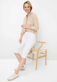 Camicia button-up in lino beige con maniche lunghe, abbinata a pantaloni bianchi fino al ginocchio e sneakers chiare, seduto su una sedia di legno.