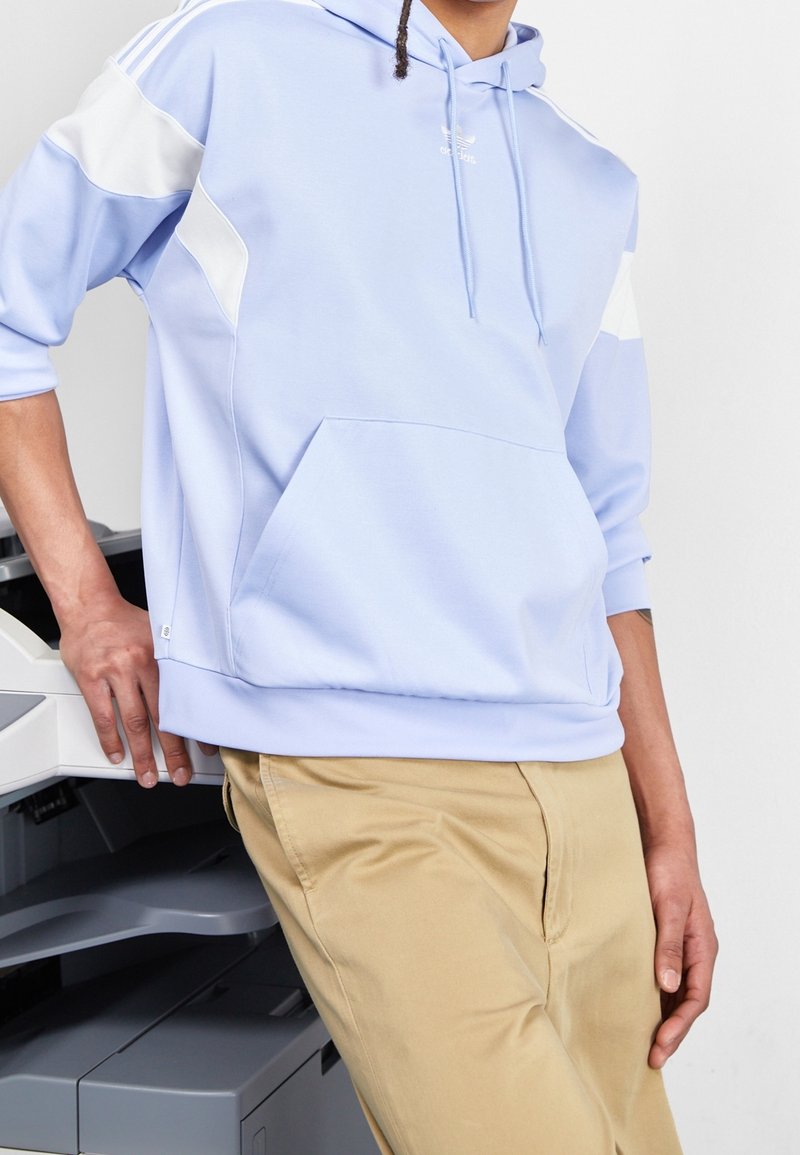 Ljusblå sweatshirt med vita detaljer, snörning och framficka. Matchas med beiga byxor, visad mot en neutral bakgrund.