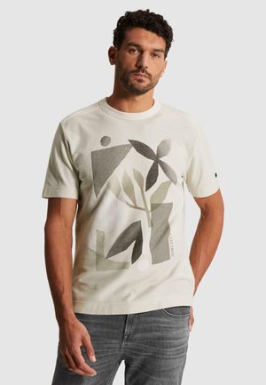 Man draagt een lichtbeige T-shirt met abstracte geometrische en bladmotieven in grijs en zwart, gecombineerd met grijze spijkerbroek.