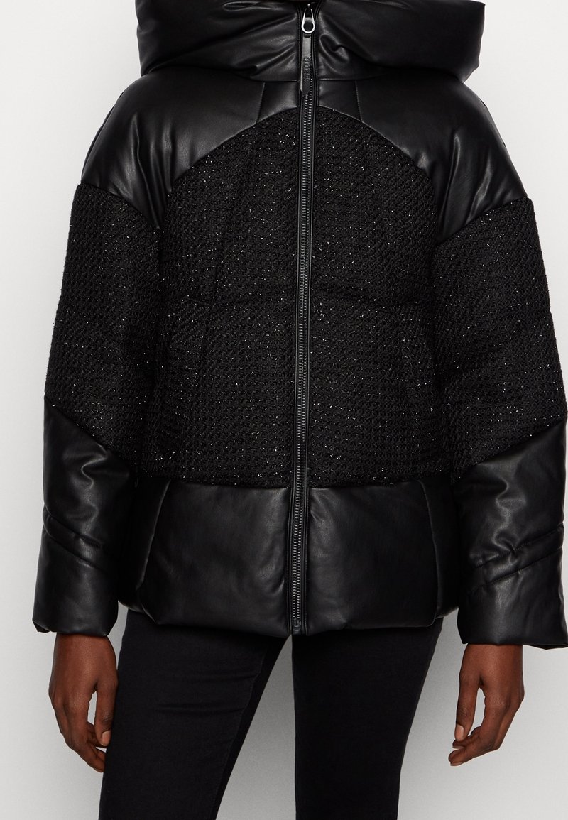 Veste matelassée noire avec des panneaux en tissu texturé, des manches en cuir et une capuche, zipée à l'avant, portée par une personne avec un pantalon foncé.