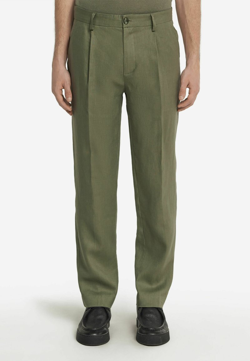 Oliwkowe, plisowane spodnie wykonane z lekkiego materiału, z zamkiem błyskawicznym i zapięciem na guzik, połączone z czarnymi butami wsuwanymi.