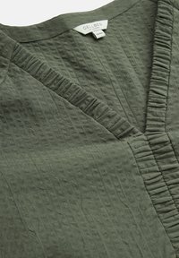 Zielona oliwkowa bluzka z teksturowanej tkaniny, z dekoltem w kształcie litery V i marszczonym akcentem wzdłuż szwu. Metka informuje "CELLBES ze Szwecji".