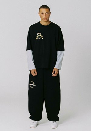 Hombre con camisa negra oversize y pantalones, con mangas largas grises debajo, de pie contra un fondo liso y claro. Zapatillas blancas visibles.