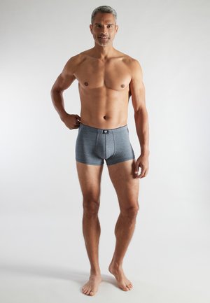 Fit middelbare man staat blootsvoets in grijze boxershorts tegen een effen witte achtergrond.
