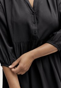 Schwarzes Tunika-Kleid mit V-Ausschnitt, Knopfleiste, gerafftem Taillenbereich und elastischen Ärmeln. Glatte Stofftextur mit fließender Silhouette.